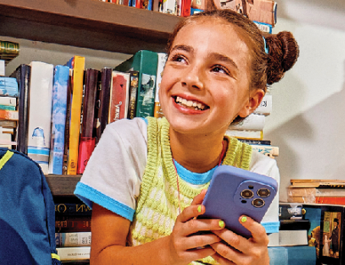 A young girl looking happily to the left while holding a smartphone and surrounded by books.