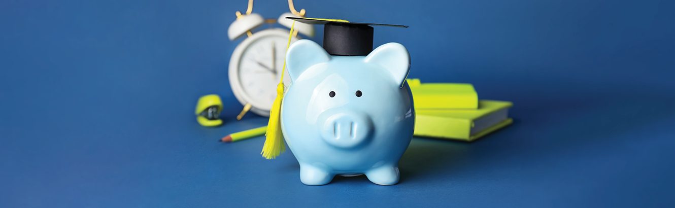 Blue piggy bank wearing a graduation cap surrounded by an alarm clock and school supplies. 